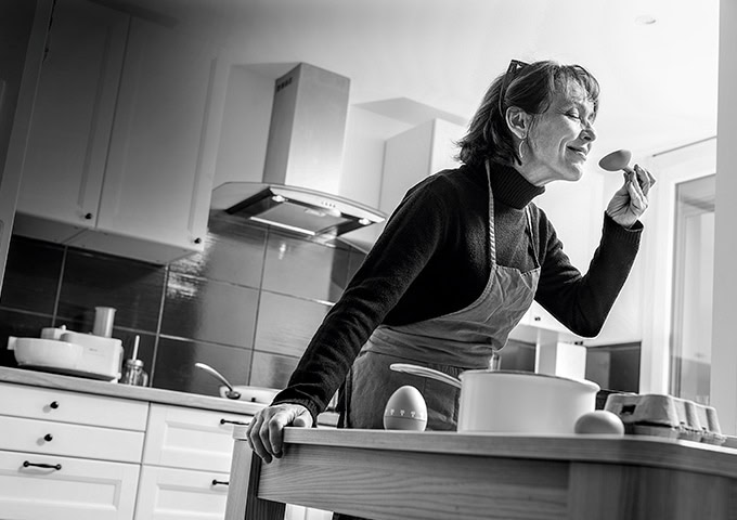 Une femme aveugle dans une cuisine. Elle sourit et tient une spatule en bois dans sa main gauche.