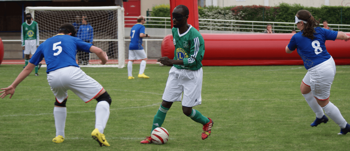 Photo d'une action d'un match de cécifoot dans lequel on voit 3 joueurs. Celui au centre est en possession du ballon et porte un maillot vert. A gauche et à droite, deux joueurs avec un maillot bleu, un homme et une femme, tentent de lui prendre le ballon.