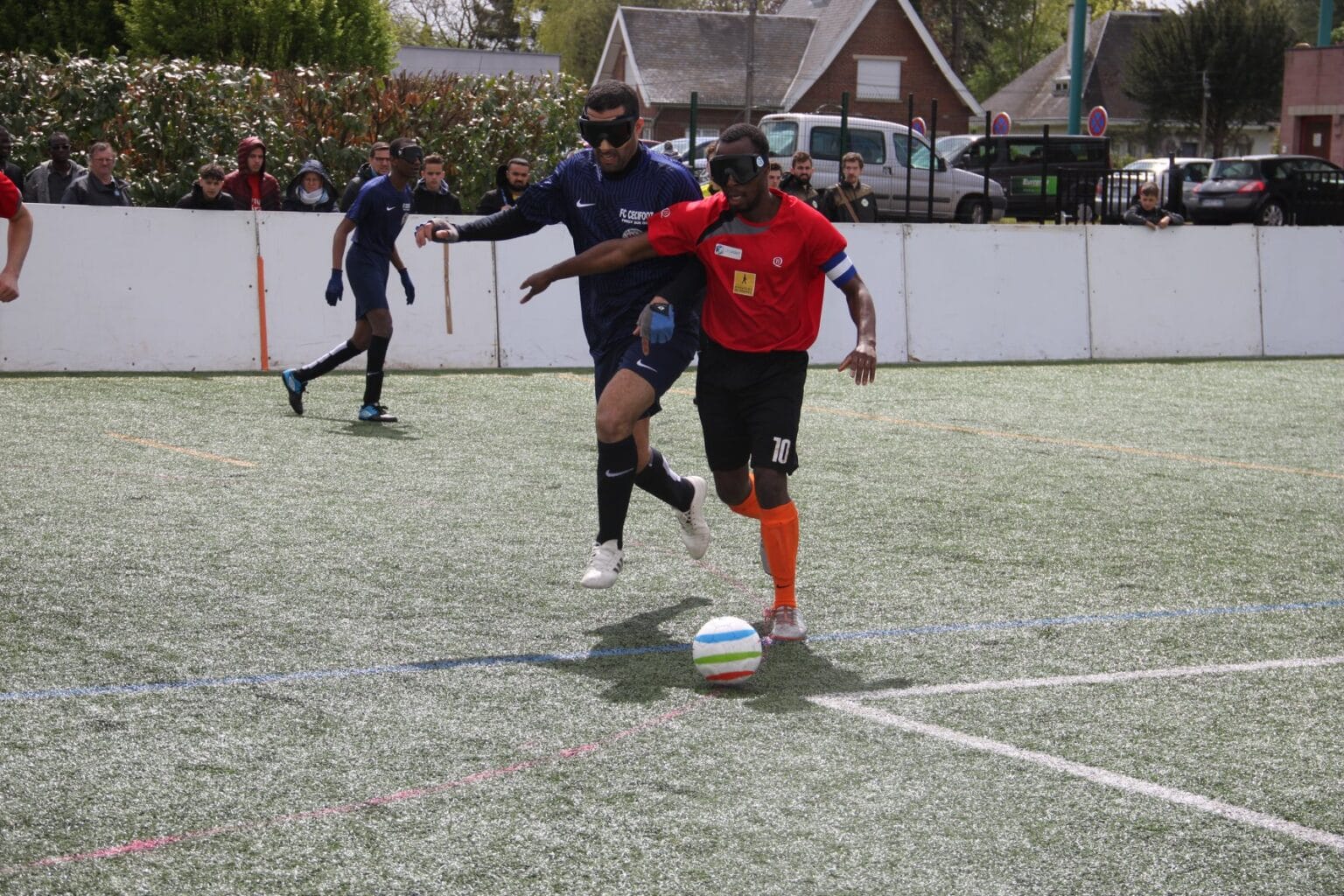 Photo de deux joueurs de cécifoot à la lutte pour le ballon. Celui avec le maillot orange est en possession du ballon et met son bras en opposition pour éviter que le joueur au maillot bleu foncé le lui prenne.