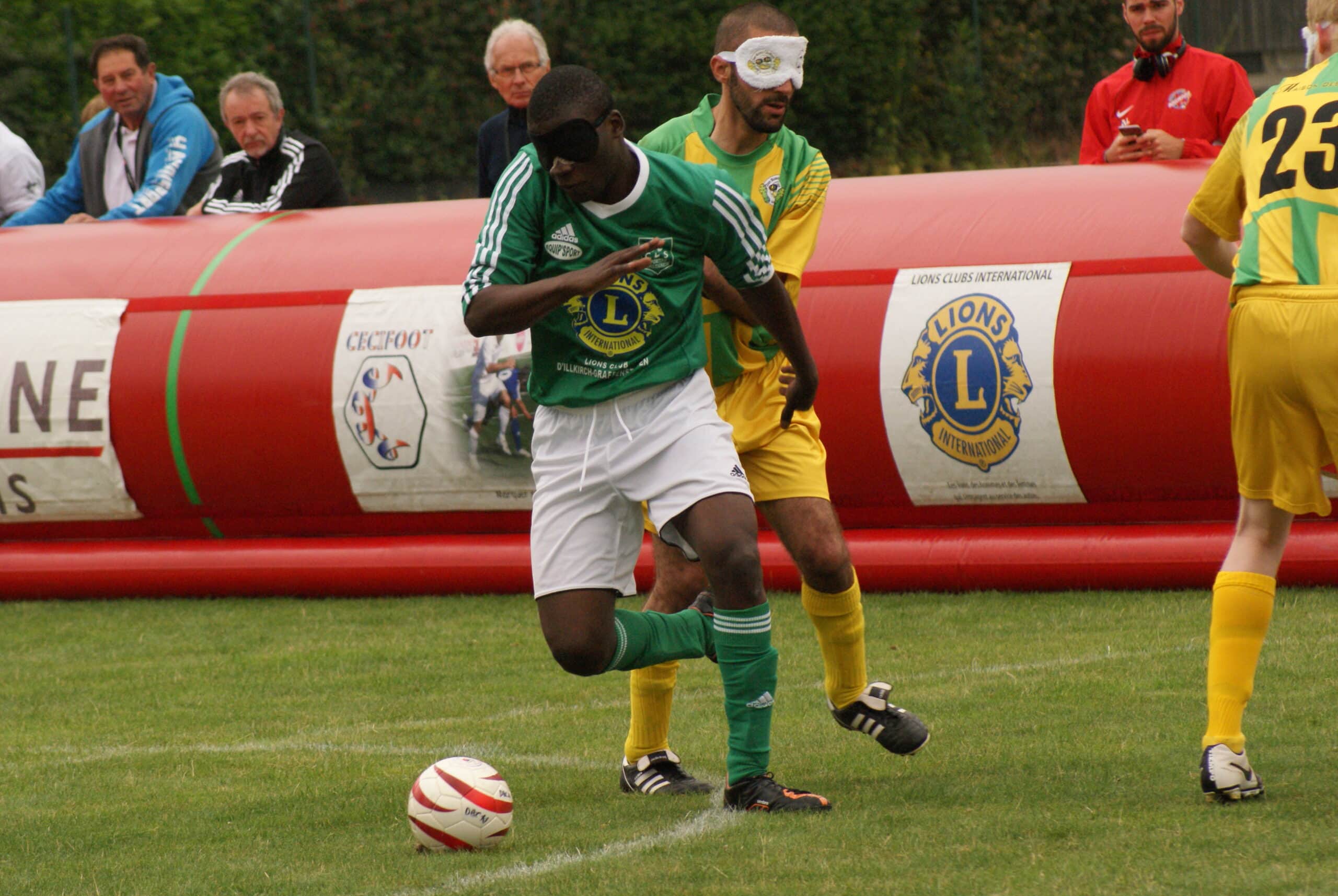 Photo d'une action d'un match de cécifoot. Un joueur au maillot vert en possession du ballon est au duel avec un joueur au maillot jaune.