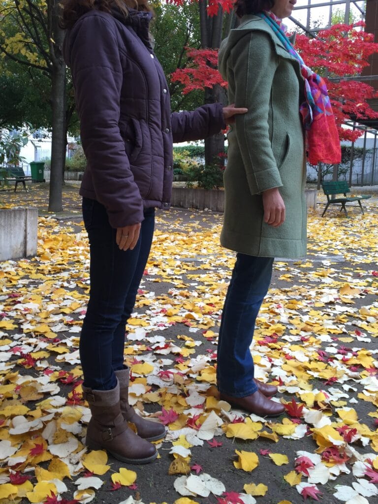 Photo de deux personnes de profil effectuant la position de base. Le guide est devant, à un pas, et garde le bras le long du corps. La personne déficiente visuelle est derrière. Elle prend le bras du guide, pouce vers l’extérieur et les autres doigts vers l’intérieur. Elle garde son coude fléchi à 90° le long de son buste.