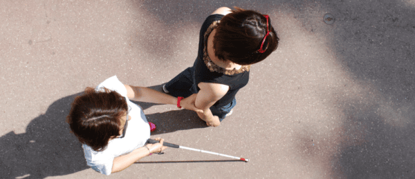 Photo vue du dessus. On voit deux femmes côtes à côtes pratiquant la technique de guide. Elles se déplacent de la gauche vers la droite de l'image. La personne guidée se situe un pas derrière sa guide, a une canne dans sa main droite et tient le coude droit de sa guide avec sa main gauche.