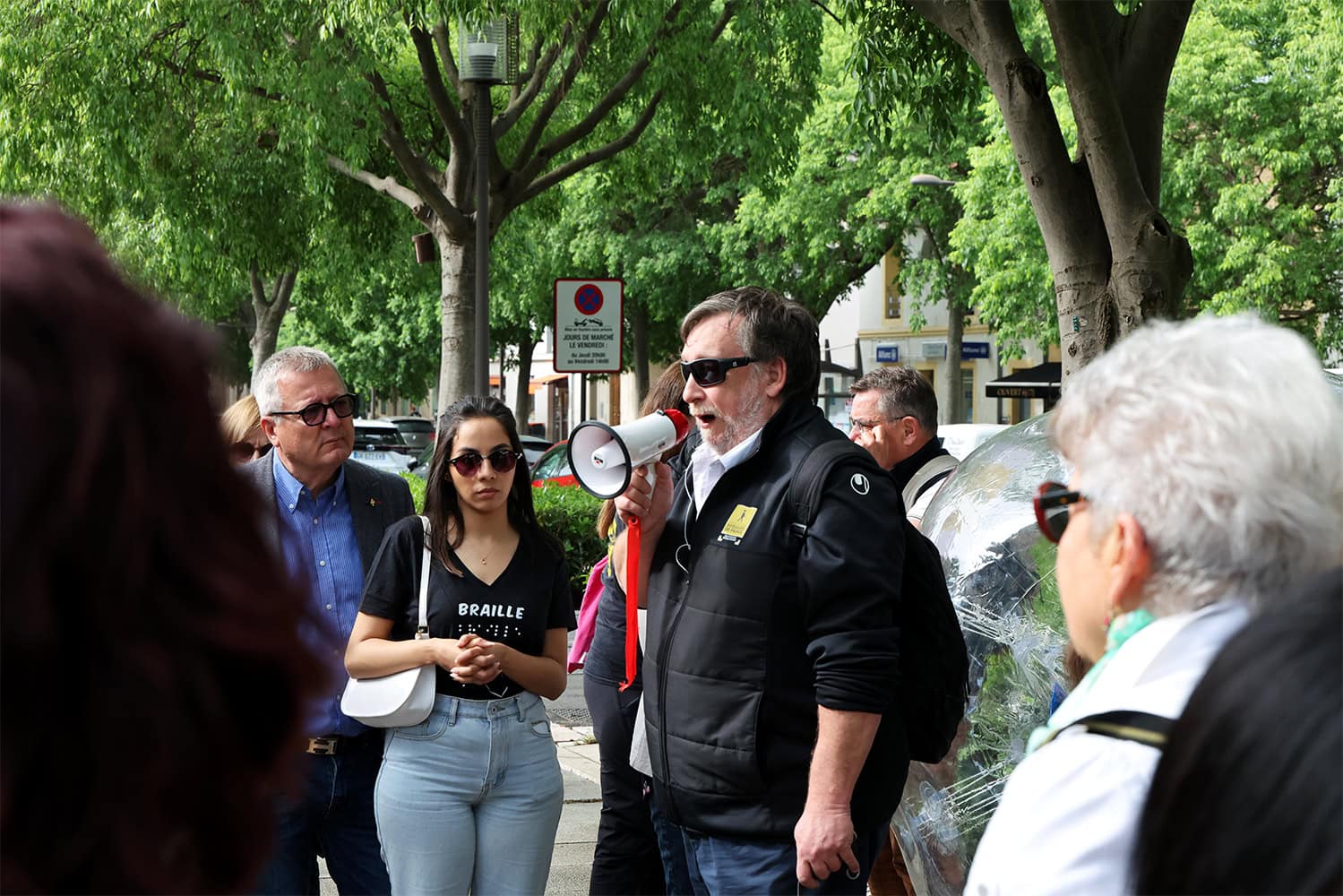 Un homme aveugle prend la parole devant un rassemblement de gens. Il tient un mégaphone dans sa main droite.