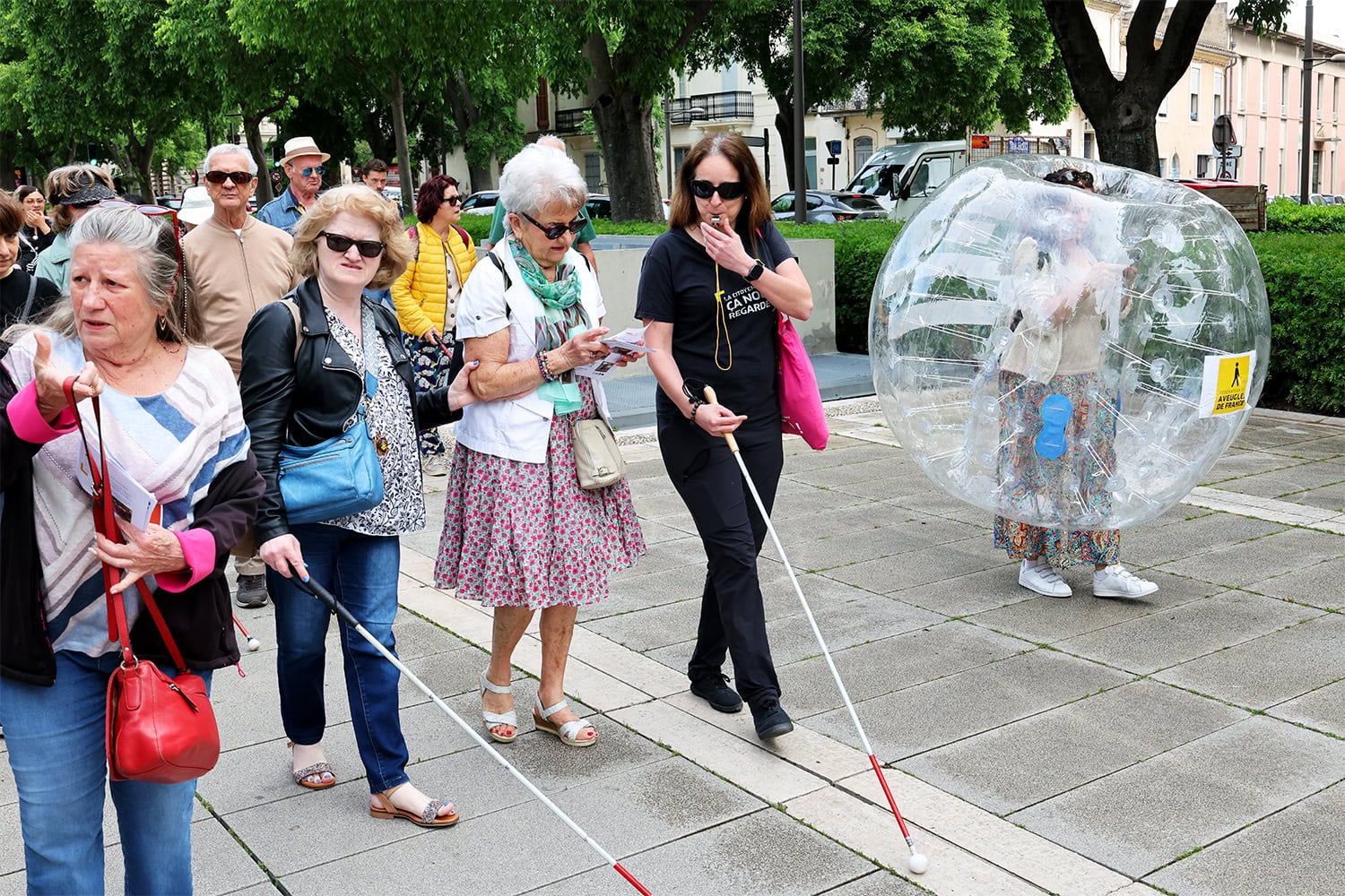 Plusieurs personnes marchent dans la rue. La plupart d'entre elles portent des cannes blanches. À droite de l'image, on voit  une grosse bulle gonflable dans laquelle une personne se tient.