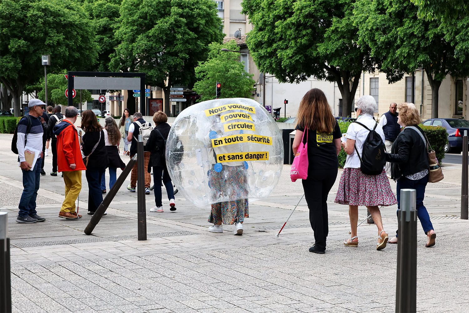 Photo de dos de plusieurs personnes qui marchent dans la rue. La plupart d'entre elles portent des cannes blanches. Au centre de l'image, on voit  une grosse bulle gonflable dans laquelle une personne se tient. Sur cette bulle est écrit le message suivant : "Nous voulons pouvoir circuler en toute sécurité".