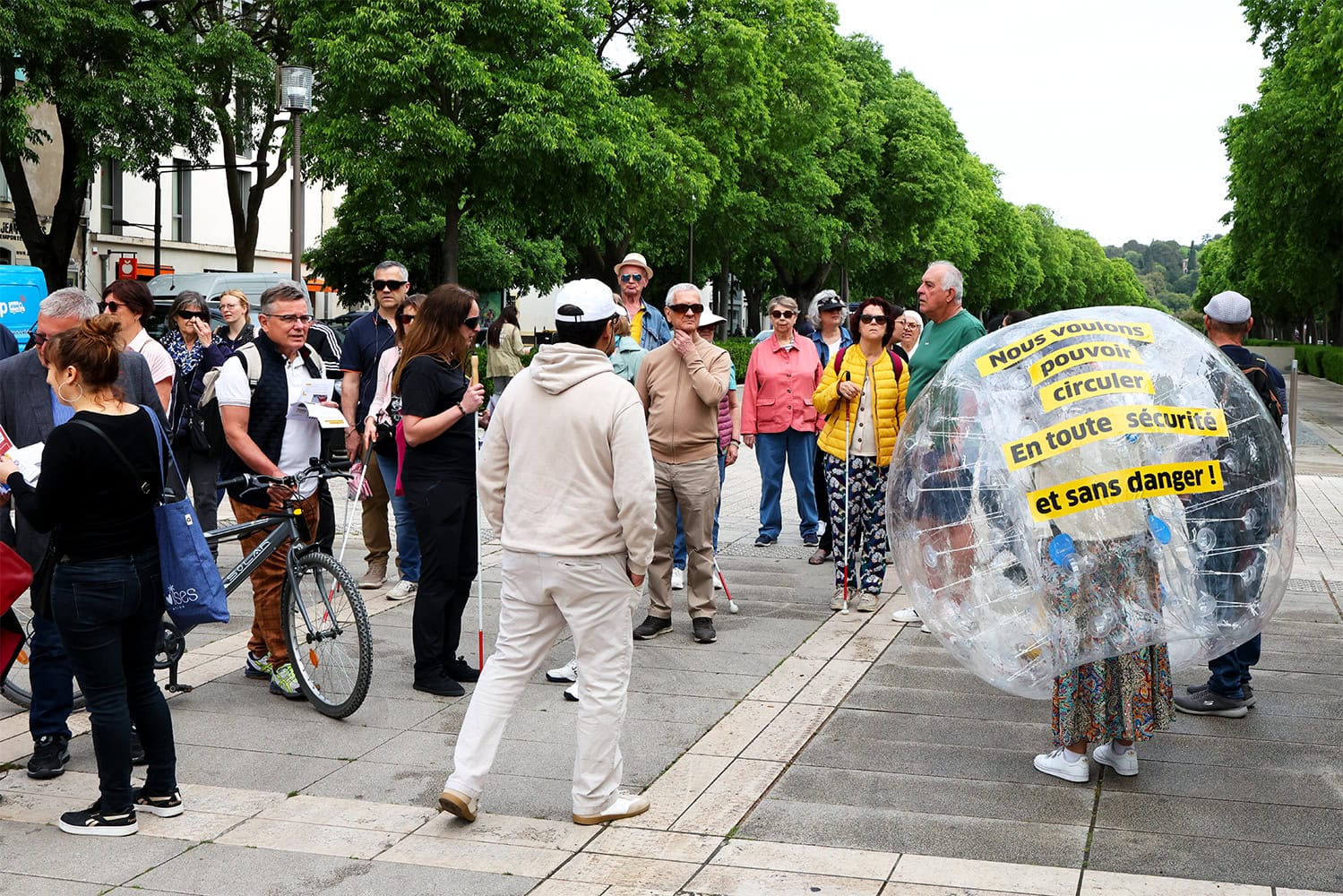 Rassemblement de dizaines de personnes sur une place. Plusieurs d'entres elles sont aveugles ou malvoyantes. Sur la droite de la photo, on voit une grosse bulle gonflable dans laquelle une personne se tient. Sur cette bulle est écrit le message suivant : "Nous voulons pouvoir circuler en toute sécurité".