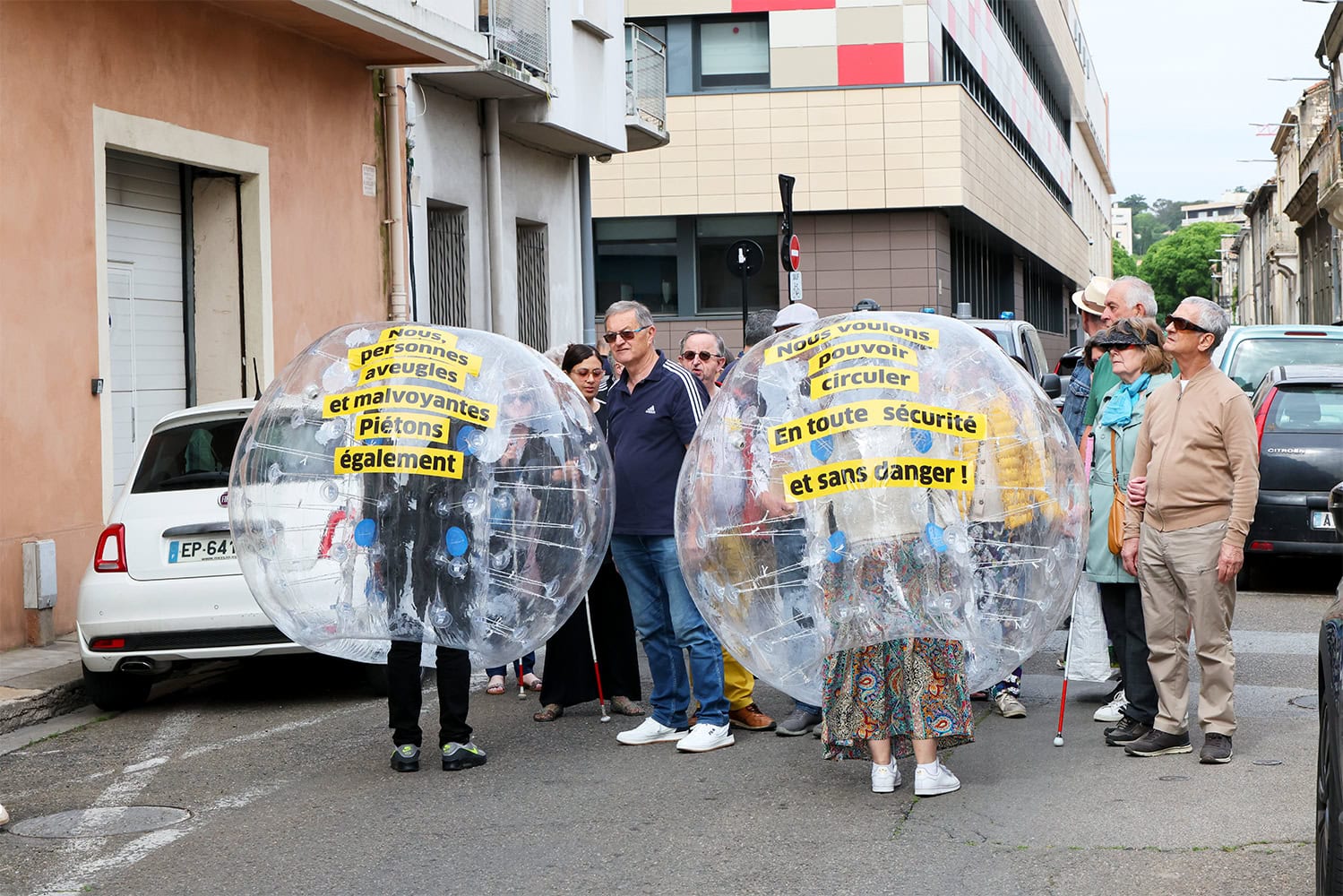 Photo de face d'un groupe de personnes déficientes visuelles. Au premier plan, on voit deux grosses bulles gonflables dans laquelle des personnes se tiennent. Sur la bulle de gauche, il est écrit le message suivant : "Nous, personnes aveugles et malvoyantes. Piétons également,". Sur la deuxième bulle, il est écrit : "Nous voulons pouvoir circuler en toute sécurité".